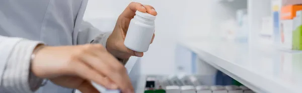 Vista recortada del farmacéutico en la botella de la celebración de la capa blanca con la medicación mientras que abre el cajón en la farmacia, bandera - foto de stock