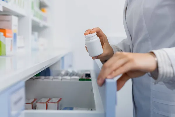 Ausgeschnittene Ansicht eines Apothekers im weißen Mantel, der eine Flasche mit Medikamenten hält, während er Schublade in der Drogerie öffnet — Stockfoto