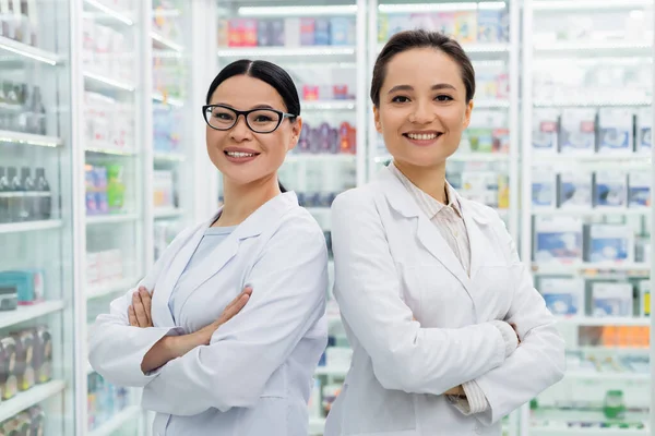 Allegri farmacisti interrazziale in cappotti bianchi in piedi con le braccia incrociate in farmacia — Foto stock