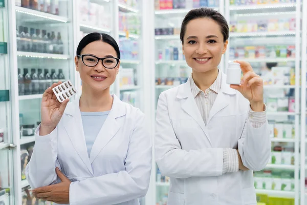 Cheerful interracial pharmacists in white coats holding medication in drugstore — Stock Photo