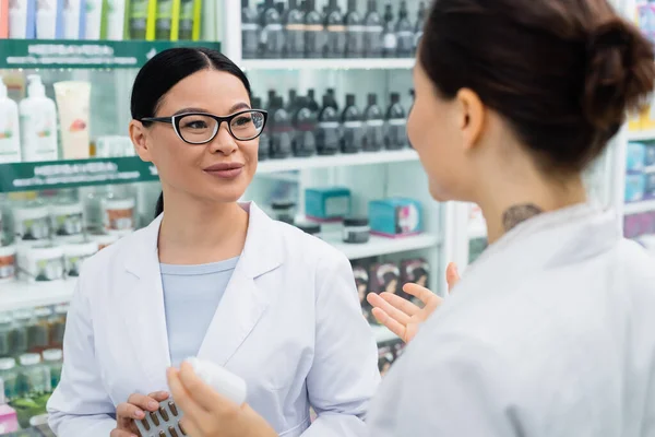 Cheerful asian pharmacist in white coat holding medication and looking at tattooed colleague — Stock Photo