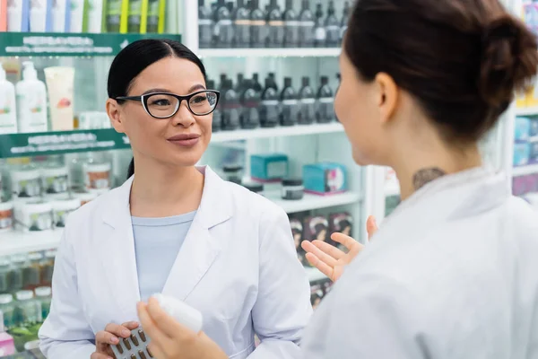 Alegre asiático farmacéutico en blanco abrigo celebración medicación y mirando tatuado colega - foto de stock