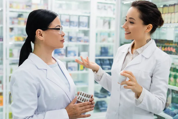 Asian pharmacist in white coat holding medication and looking at tattooed colleague — Stock Photo