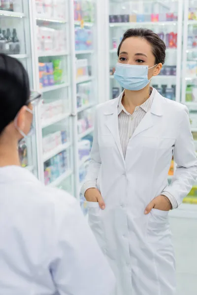 Farmacéutico tatuado en máscara médica y abrigo blanco de pie con las manos en los bolsillos cerca de colega borrosa - foto de stock