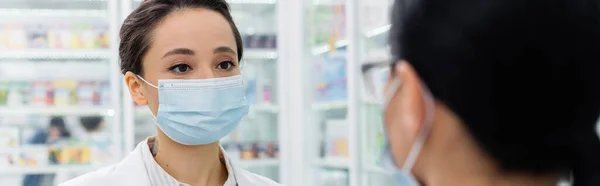 Pharmacien tatoué dans un masque médical en regardant un collègue flou, bannière — Photo de stock