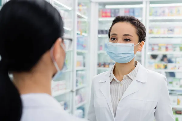 Farmacéutico tatuado en máscara médica mirando colega borrosa - foto de stock