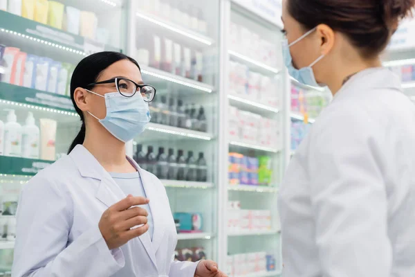 Asiatischer Apotheker in medizinischer Maske blickt verschwommenen Kollegen an — Stockfoto