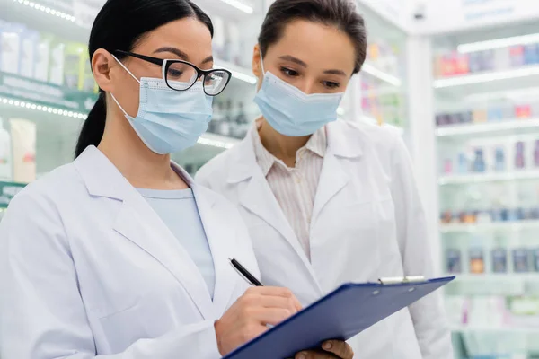 Asian pharmacist in medical mask writing on clipboard near colleague — Stock Photo