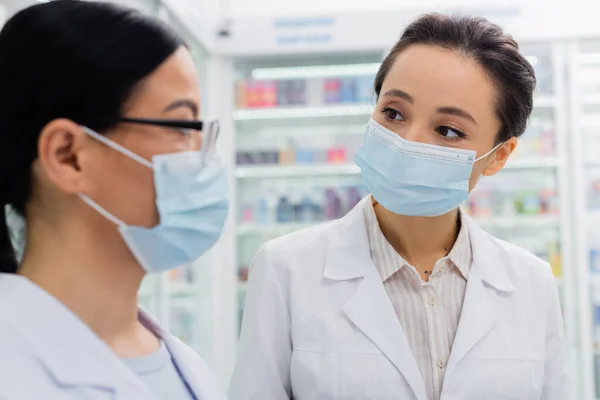 Farmacéuticos interraciales con máscaras médicas mirándose en la farmacia - foto de stock