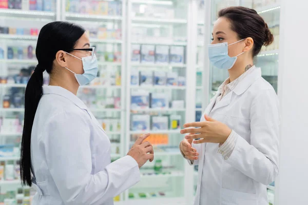 Interrassische Apotheker in medizinischen Masken schauen einander beim Gespräch in der Drogerie an — Stockfoto