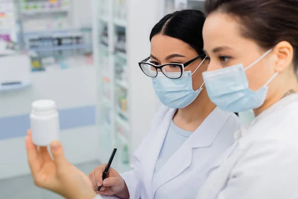 Interracial pharmacists in medical masks looking at bottle in drugstore — Stock Photo