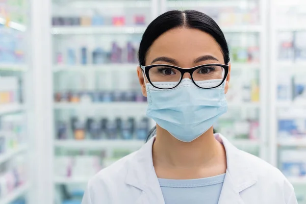 Asiático farmacéutico en gafas y médico máscara mirando a cámara - foto de stock