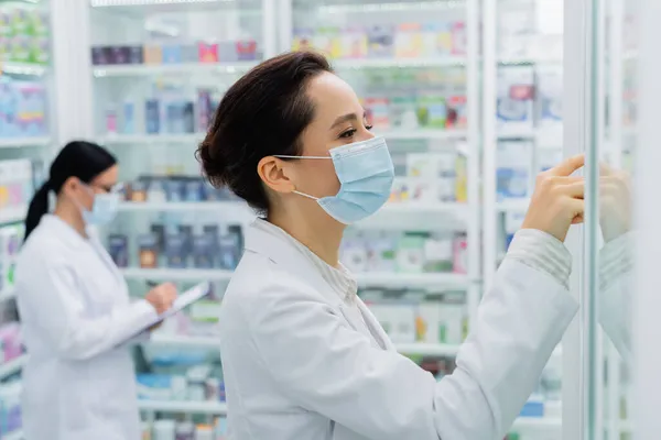 Farmacista in maschera medica che controlla i farmaci vicino al collega offuscato — Foto stock