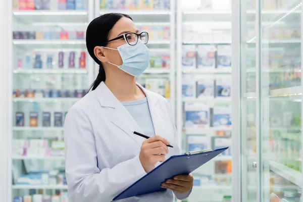 Asiatischer Apotheker in Brille und medizinischer Maske mit Klemmbrett bei der Medikamentenkontrolle — Stockfoto