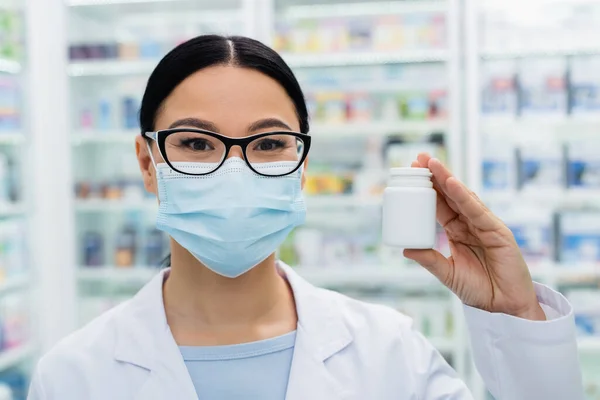 Asiático farmacéutico en gafas y médico máscara celebración botella con pastillas - foto de stock