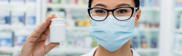 Азіатський фармацевт в окулярах і медична маска тримає пляшку з таблетками, банер — стокове фото