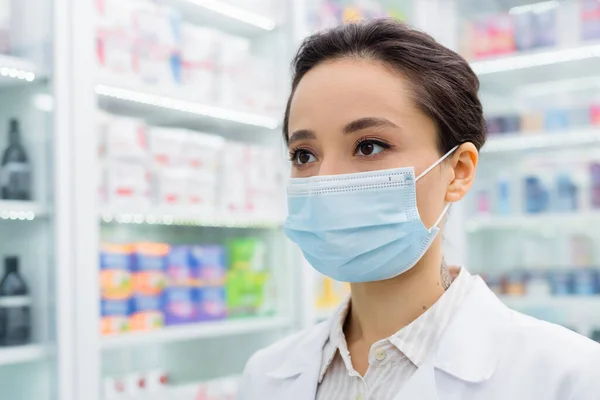 Boticario tatuado en máscara médica mirando hacia otro lado en la farmacia - foto de stock