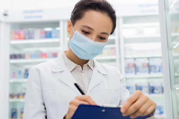 Brunette apothecary in medical mask writing on clipboard — Stock Photo