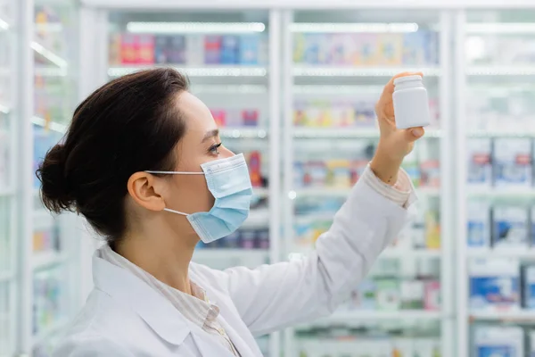 Vista laterale del farmacista bruna in maschera medica guardando bottiglia in farmacia — Foto stock