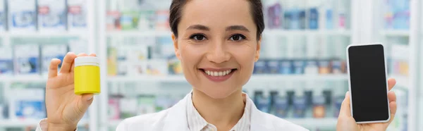 Farmacéutico alegre en la capa blanca que sostiene el teléfono inteligente con la pantalla en blanco y la botella en farmacia — Stock Photo