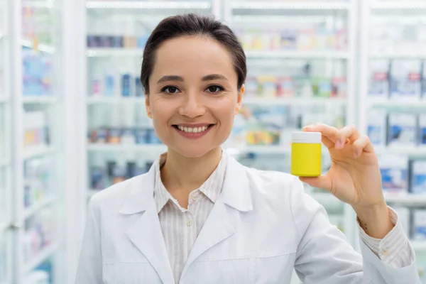 Farmacéutico alegre en la botella blanca de la celebración de la capa en la farmacia - foto de stock