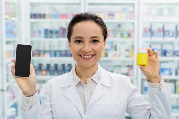 Pharmacien gai en manteau blanc tenant smartphone avec écran vierge et bouteille en pharmacie — Photo de stock