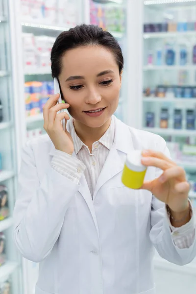 Allegro farmacista in cappotto bianco che parla su smartphone e tiene la bottiglia in farmacia — Foto stock