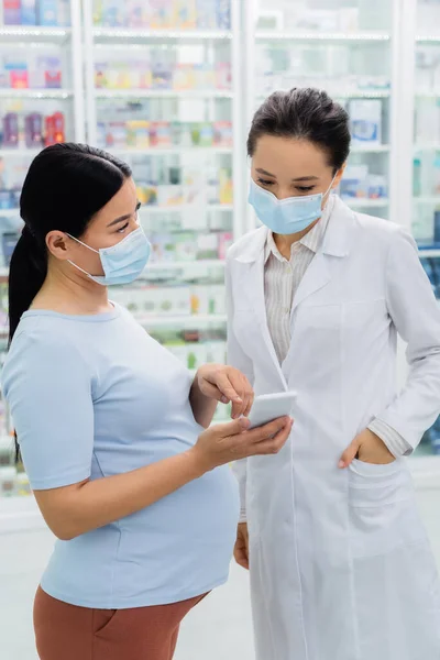 Asian pregnant woman in medical mask pointing at smartphone near pharmacist in drugstore — Stock Photo