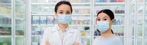 Farmacéutico en médico máscara de pie cerca asiático cliente en droguería, bandera - foto de stock