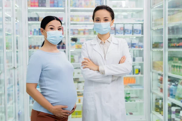 Farmacéutico en médico máscara de pie con los brazos cruzados cerca asiático embarazada mujer en droguería - foto de stock