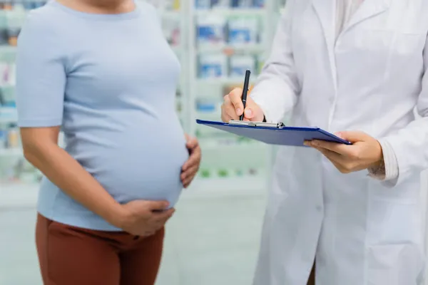 Vue recadrée de la femme enceinte debout près du pharmacien écrivant sur presse-papiers — Photo de stock