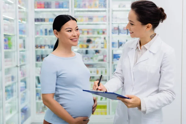 Asiatische schwangere Frau lächelt, während sie tätowierte Apothekenschrift auf Klemmbrett betrachtet — Stockfoto