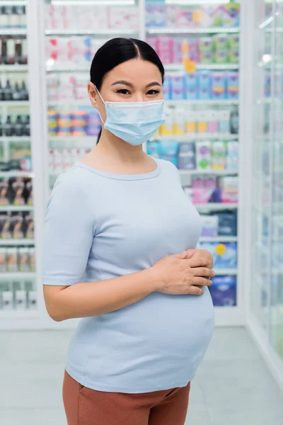 Беременный азиатский клиент в медицинской маске смотрит в камеру в аптеке — стоковое фото