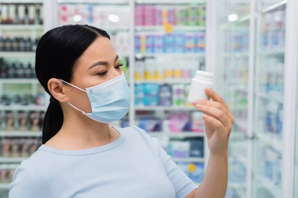 Mujer asiática en máscara médica mirando botella con vitaminas en farmacia - foto de stock