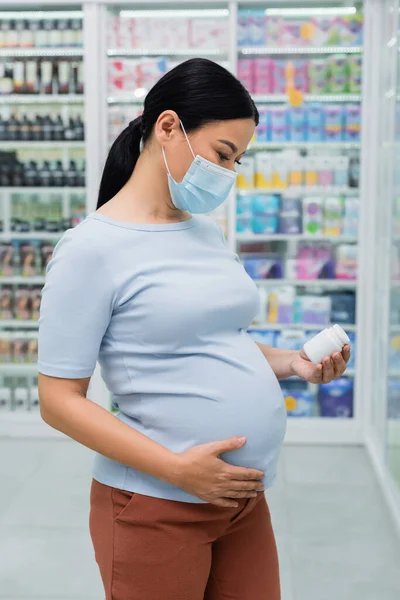 Embarazada asiático cliente en médico máscara buscando en botella con vitaminas en droguería - foto de stock