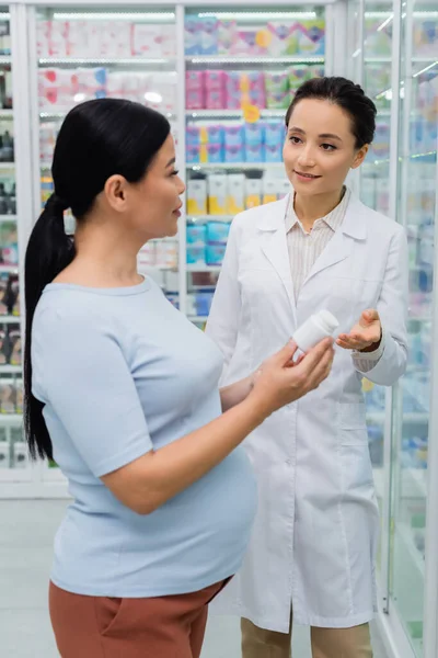 Фармацевт указывает на бутылку в руках беременной азиатки в аптеке — стоковое фото