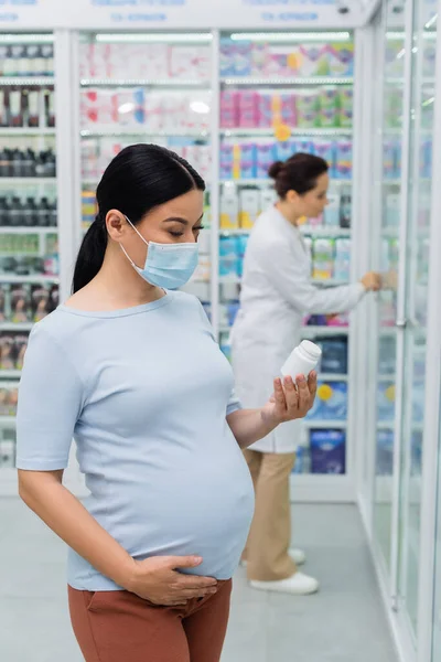 Embarazada asiático mujer en médico máscara mirando en botella en droguería - foto de stock