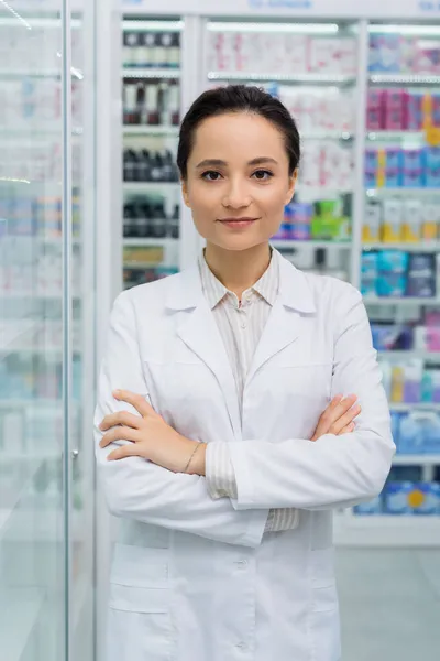 Pharmacien brunette regardant la caméra debout avec les bras croisés dans la pharmacie — Photo de stock