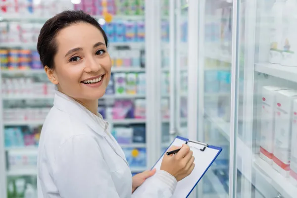 Joyful pharmacist looking at camera writing on clipboard — Stock Photo