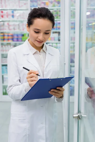Cheerful pharmacist in white coat writing on clipboard — Stock Photo