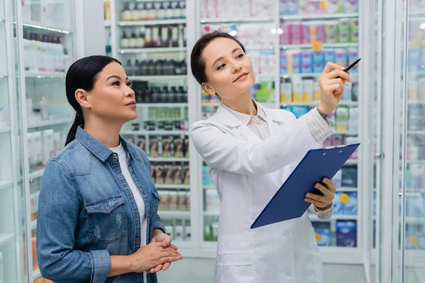 Farmacêutico apontando com caneta enquanto segurando prancheta perto de mulher asiática na farmácia — Fotografia de Stock