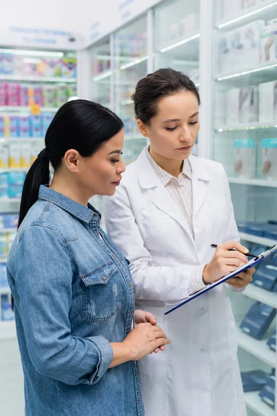 Farmacéutico escritura en portapapeles cerca asiático mujer en droguería - foto de stock