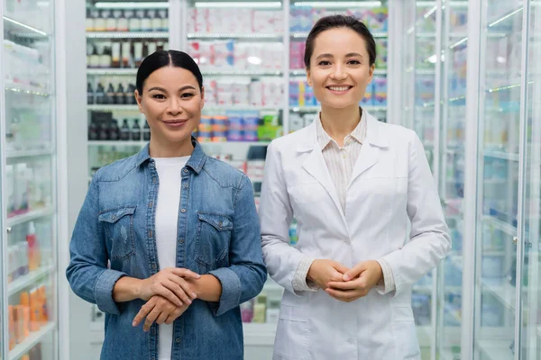 Happy pharmacist in white coat and smiling asian woman in drugstore — Stock Photo