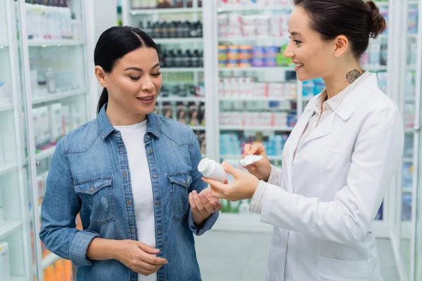 Farmacista tatuato suggerendo farmaci per sorridere donna asiatica in farmacia — Foto stock