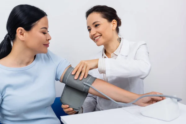 Tattooed doctor in white coat smiling while adjusting tonometer on asian patient — Stock Photo