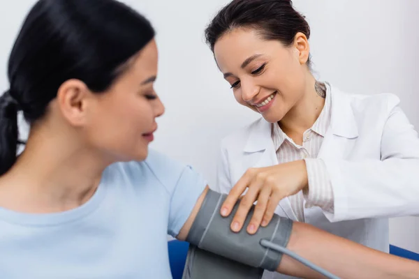 Tattooed doctor in white coat smiling while adjusting tonometer on blurred asian patient — Stock Photo