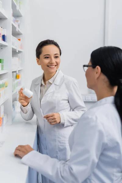 Веселый фармацевт в белом халате держит бутылку с лекарствами рядом с размытым азиатским коллегой в очках — стоковое фото