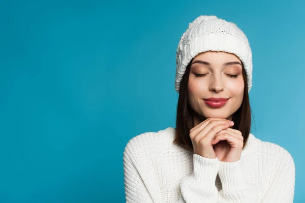 Mujer joven en suéter y sombrero de pie con los ojos cerrados aislados en azul - foto de stock