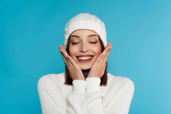 Cheerful woman in warm hat and sweater touching cheeks isolated on blue — Stock Photo