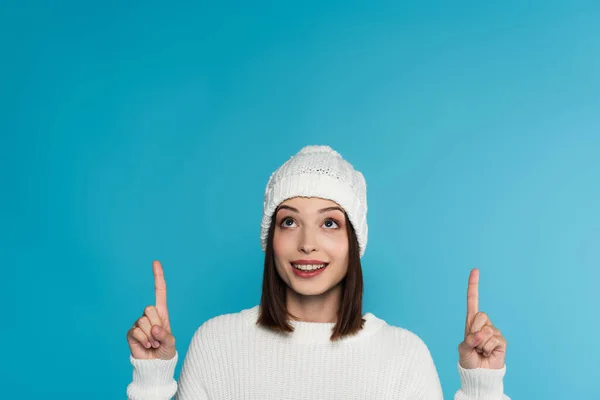 Mujer sonriente en suéter cálido y sombrero apuntando con los dedos hacia arriba aislado en azul - foto de stock